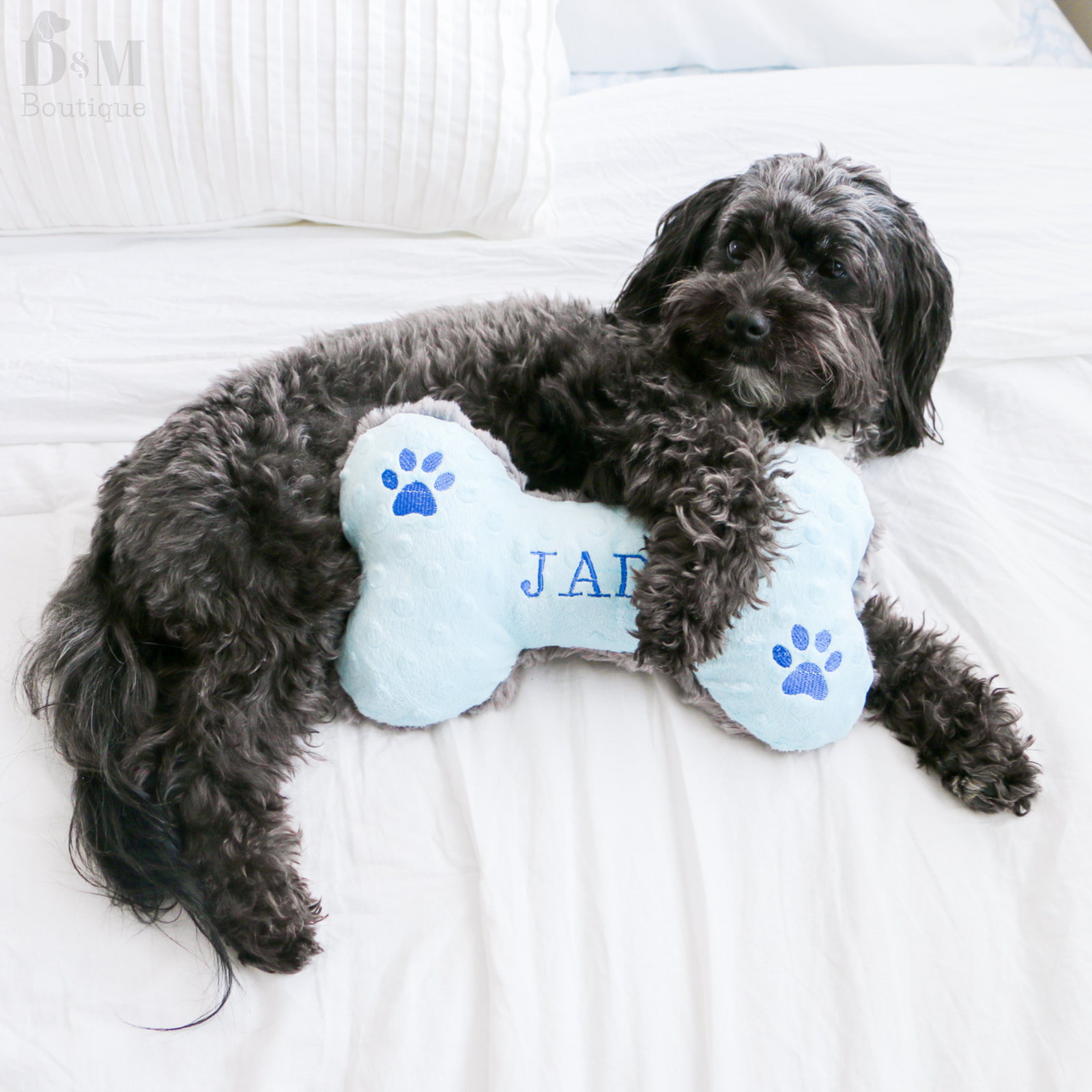 Dog lying on a white bed with a bone-shaped toy that has 'JAX' embroidered on it.