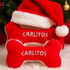 Red bone-shaped pet toy with Santa hat and 'CARLITOS' text, against a blurred Christmas tree background.