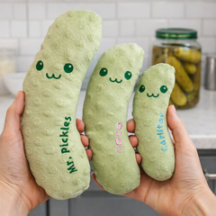 Three plush pickle toys with names embroidered on them, held in front of a kitchen background.
