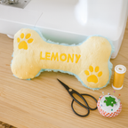 Bone-shaped plush toy with 'LEMONY' on a sewing table with scissors and thread spool.