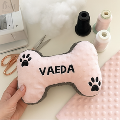 Pink bone-shaped toy with 'VAEDA' branding held by a hand, on a surface with sewing materials.