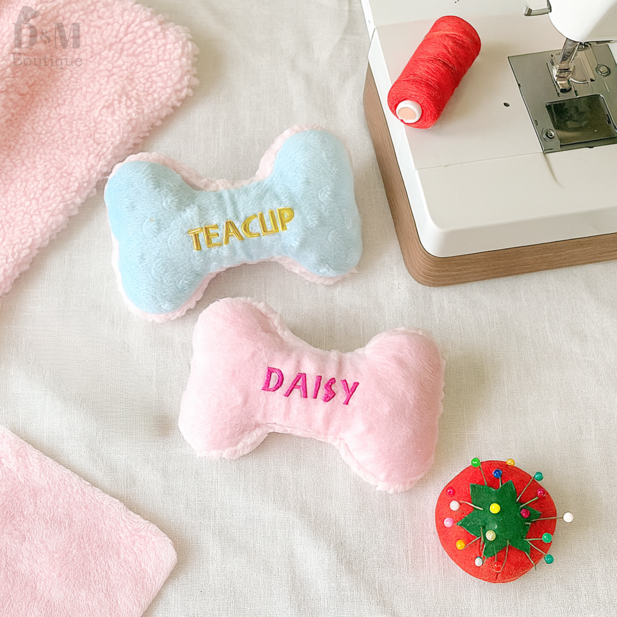 Pink and blue bone-shaped plush toys with embroidered names on a sewing machine table.