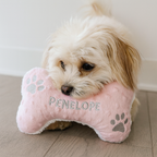Small dog with a pink bone-shaped pillow named 'Penelope' on a wooden floor.