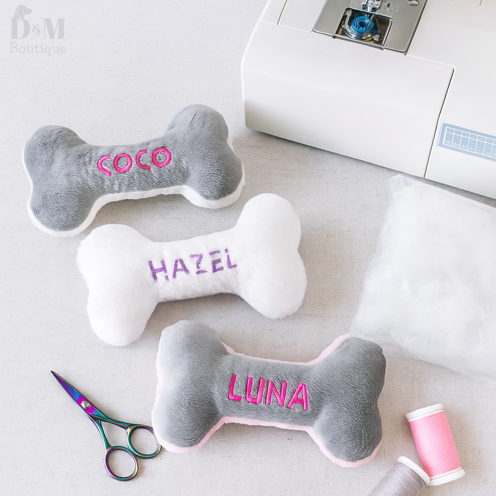 Three bone-shaped dog toys with embroidered names on a table with sewing equipment.