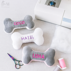 Three bone-shaped dog toys with embroidered names on a table with sewing equipment.